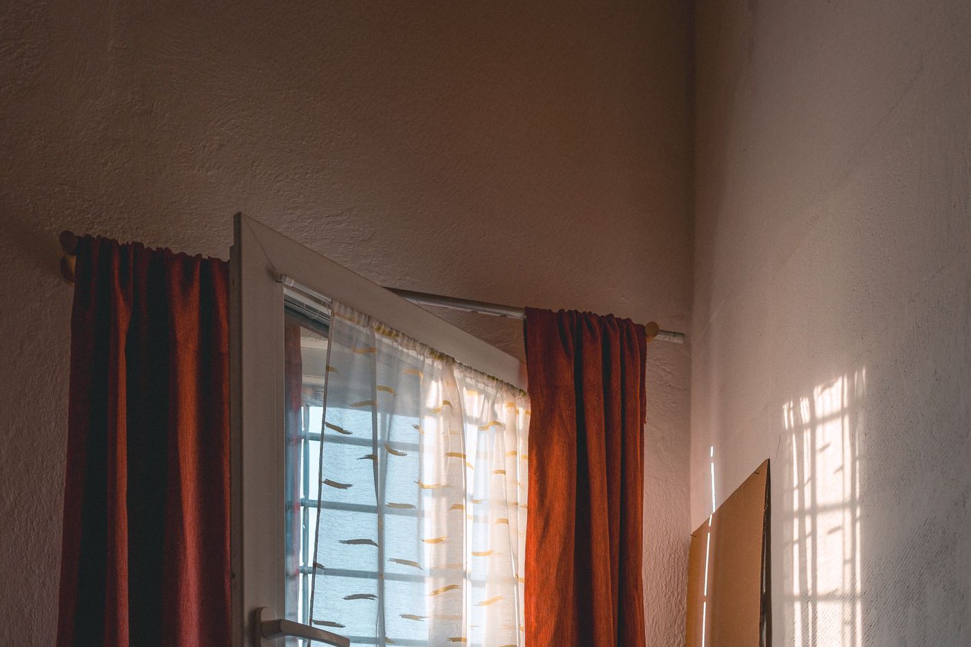 Red curtains gathered on a slim metal rod mounted above an open white window in a white-walled room.