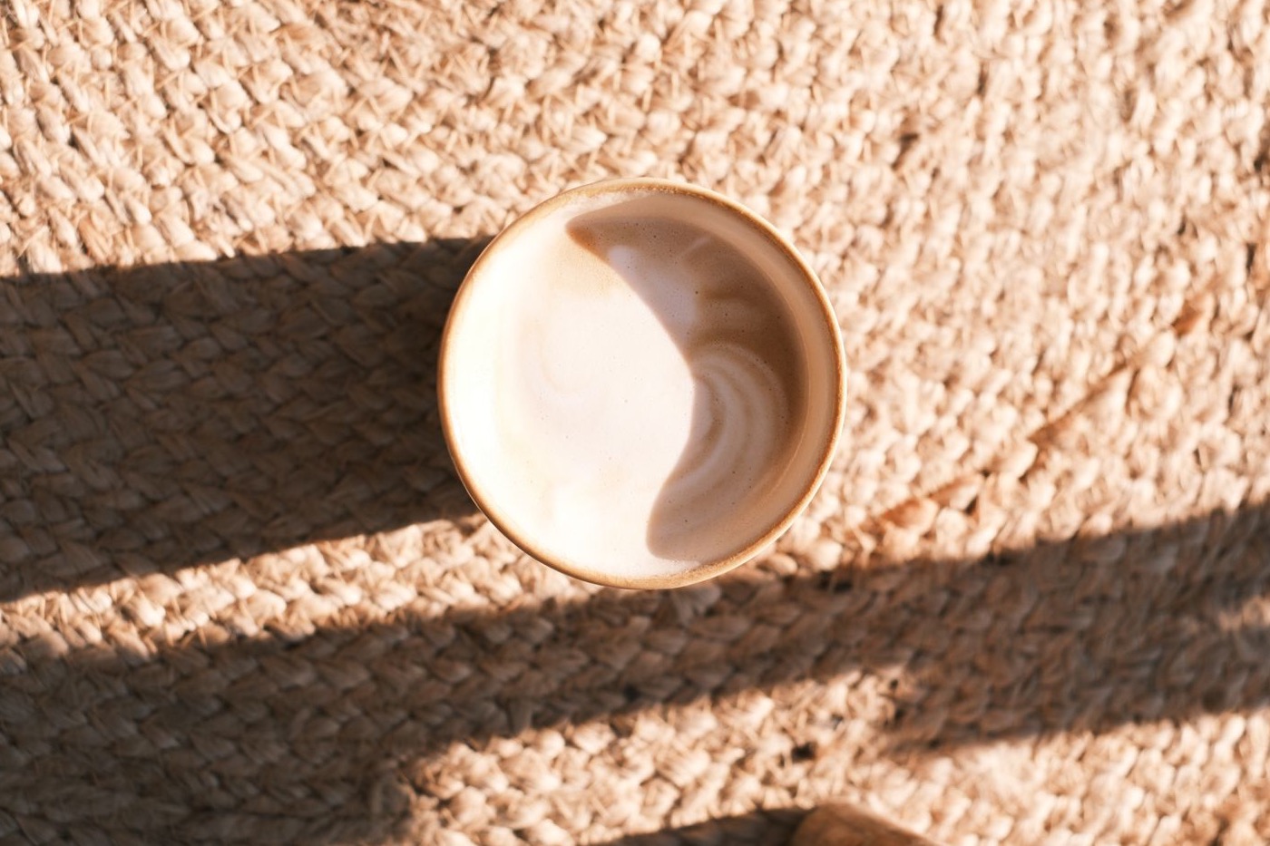 A cup of coffee sitting on a textured woven jute rug in warm sunlight, casting a long shadow across the pile.